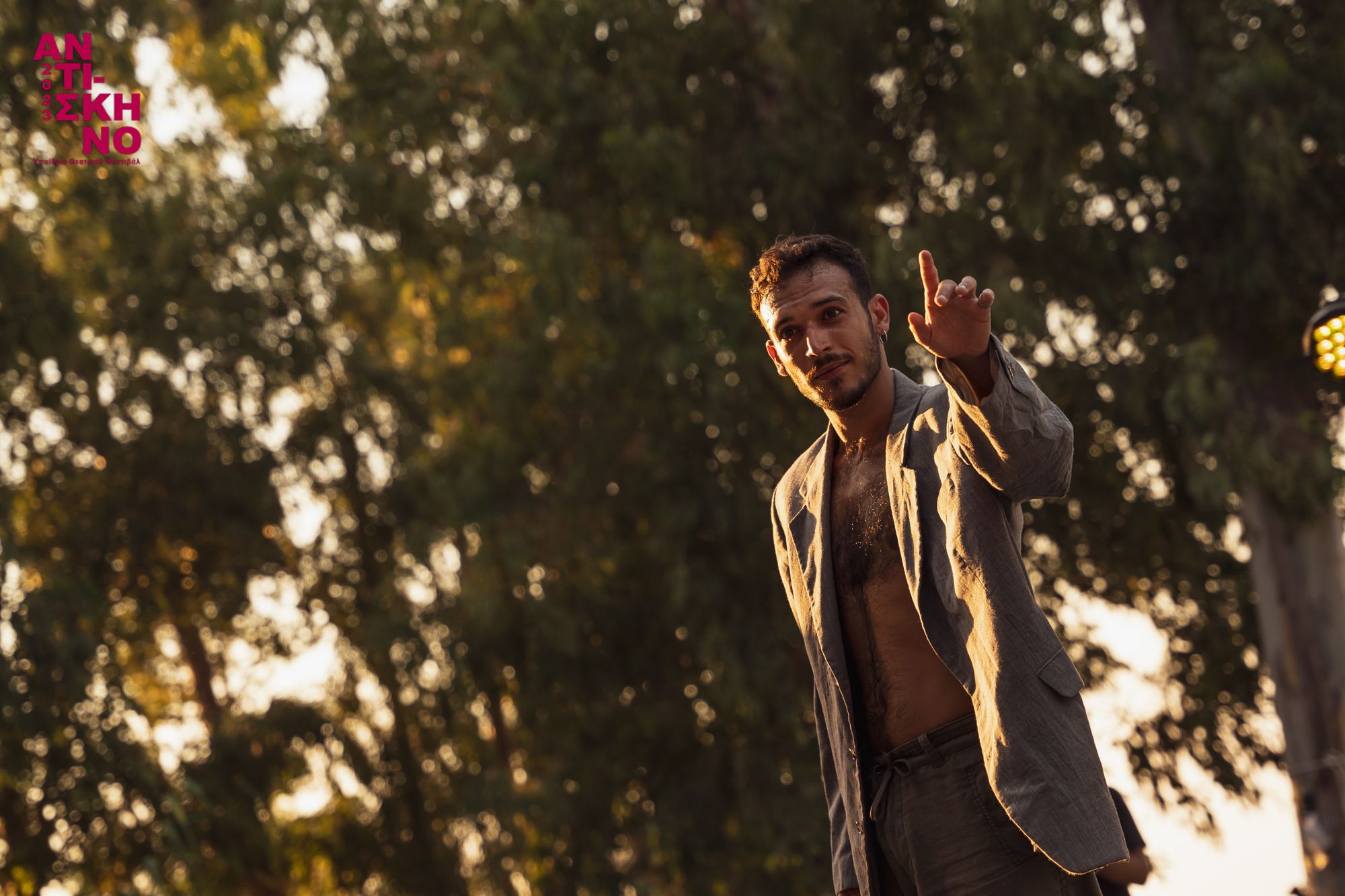 Alexandros pointing toward the audience on an outdoor stage among trees