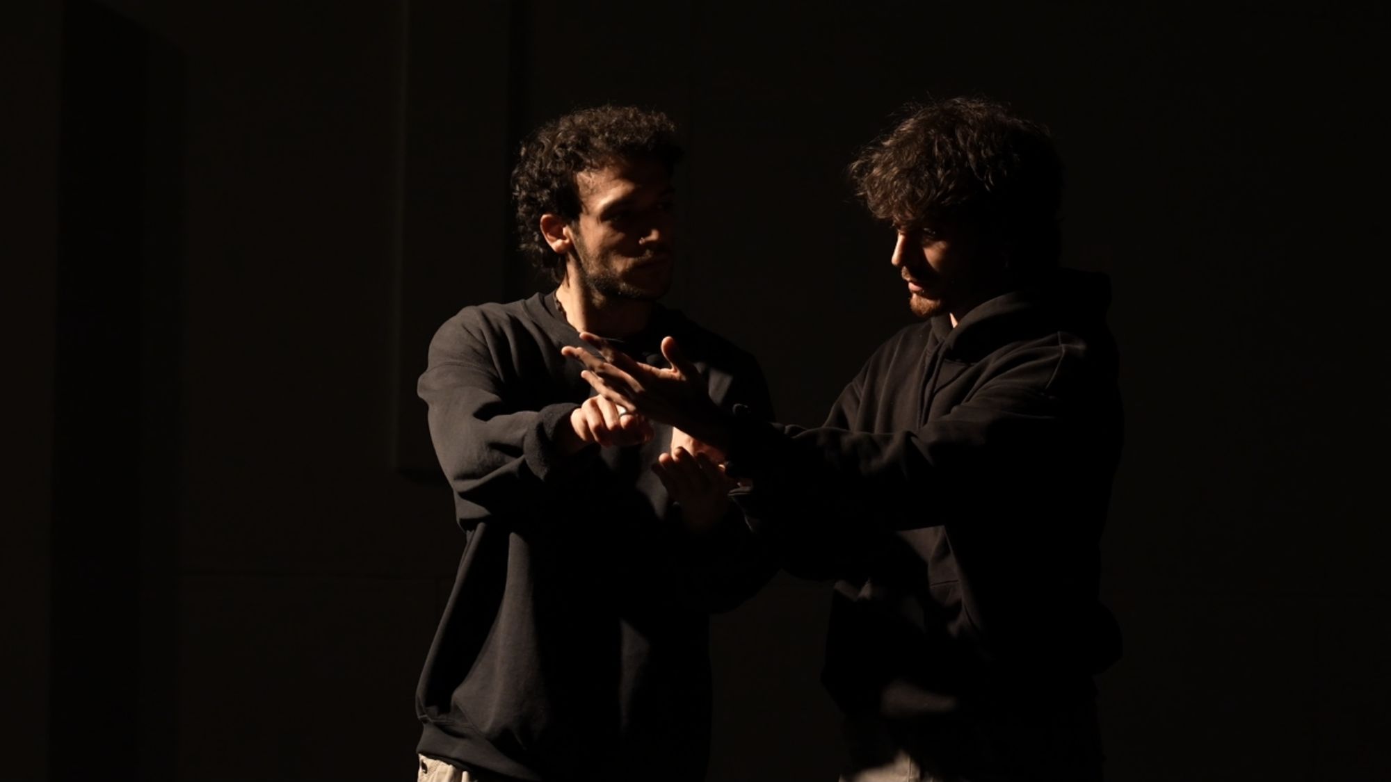 The two performers in dark jumpers facing each other, hands raised, on a dark stage