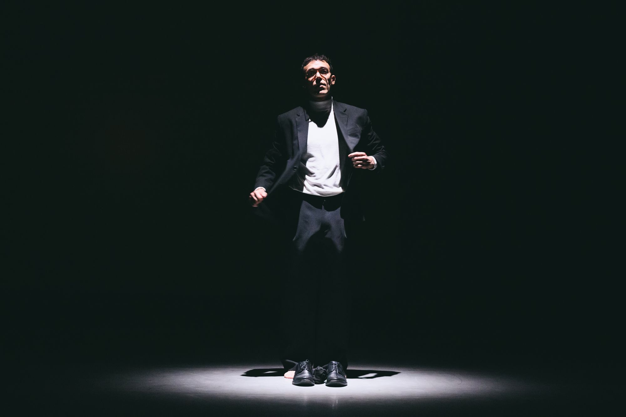 Alexandros in a dark suit standing in a pool of light