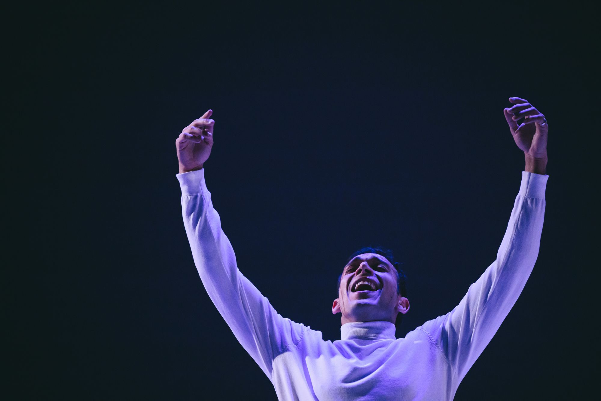 Alexandros, arms raised and laughing, in a white turtleneck under blue stage light