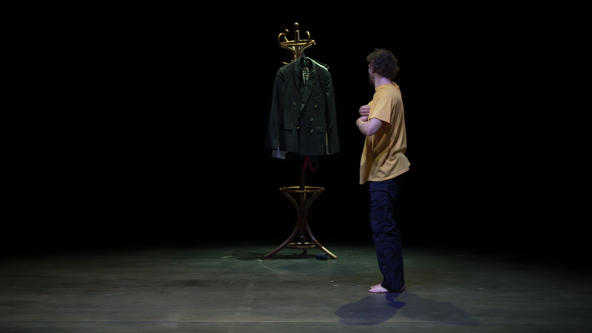 A performer facing a coat rack with a green jacket, alone in a pool of light