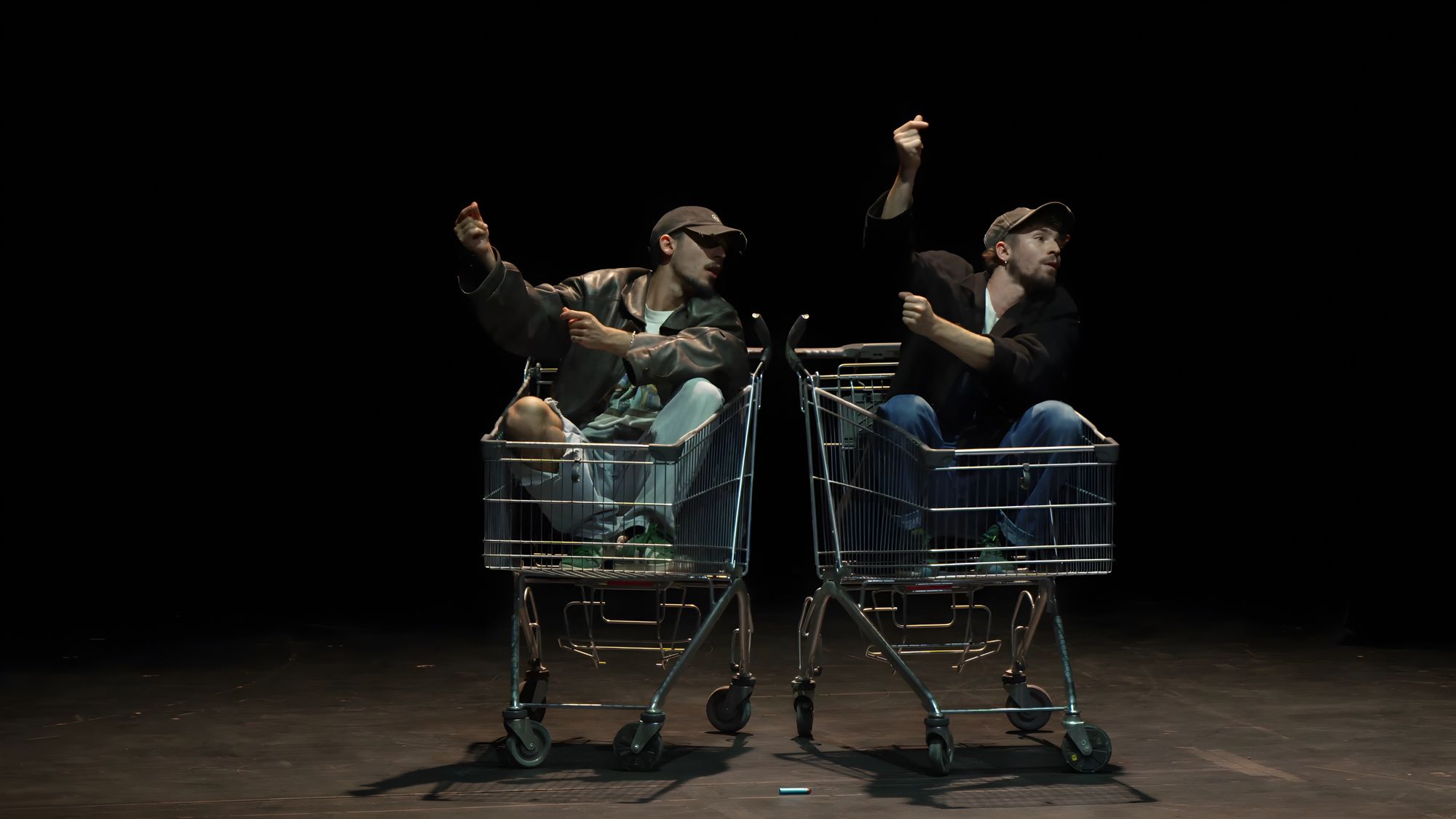 Two performers in caps sitting in shopping carts on a dark stage